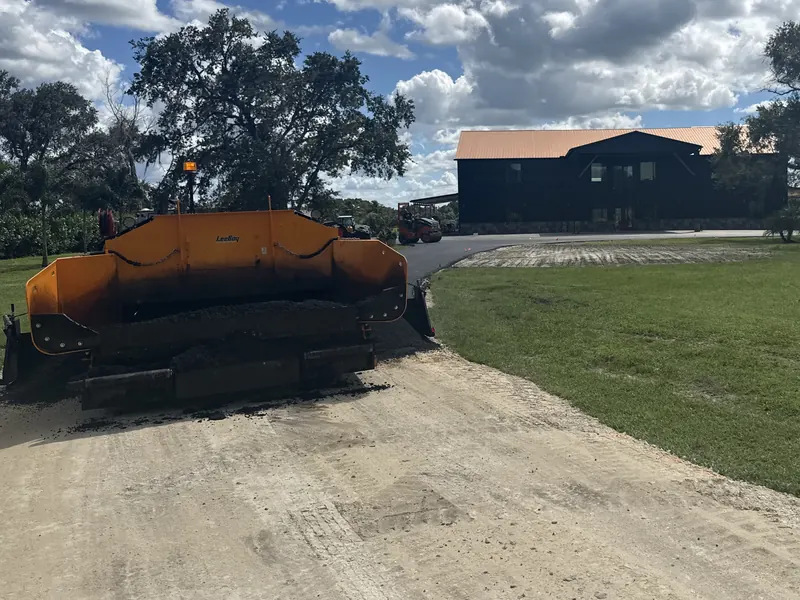 Asphalt paver working on a prepared sand base beside a lawn with live oaks