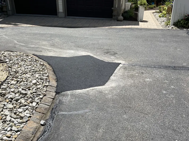 Fresh asphalt patch cut into a worn driveway beside a paver border