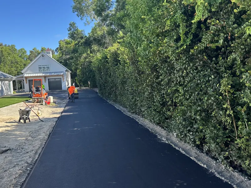 Fresh asphalt driveway leading to a white Florida cottage, crew finishing at the garage